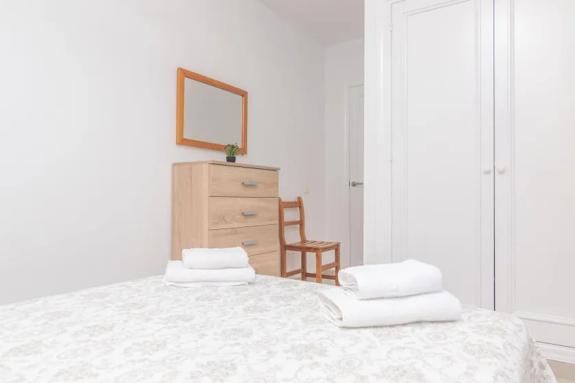 A bright bedroom with a dresser, mirror, and folded linens on the bed.