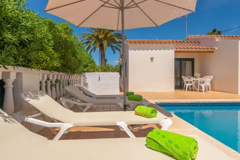 A sunny poolside patio with lounge seating, green towels, and a shaded outdoor dining area.
