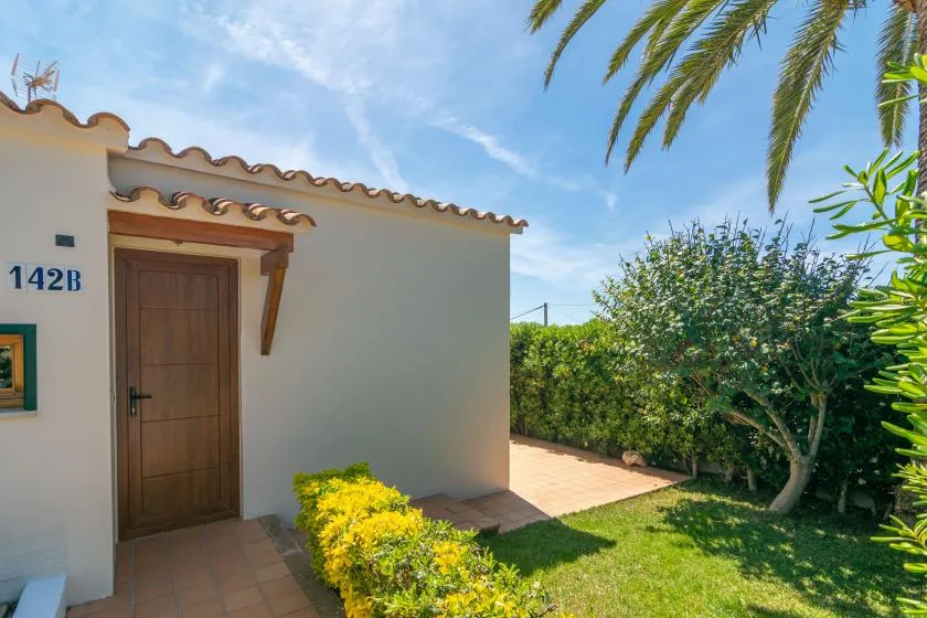 A white exterior wall and wooden entrance door beside a sunny garden.