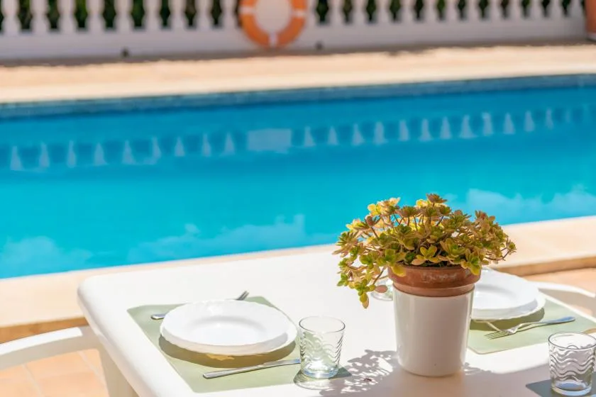 A poolside dining table is set with plates and a potted plant.
