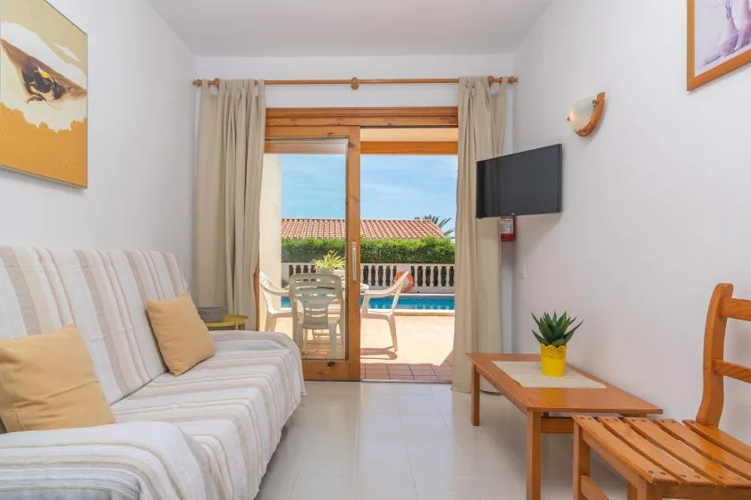 A bright living room opens onto a balcony, with seating and a wall-mounted television.