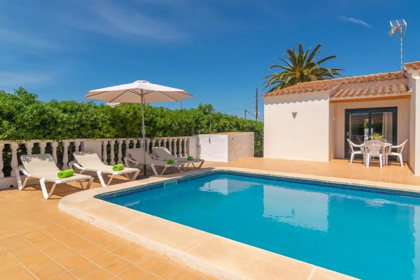 A swimming pool beside a tiled patio, sun loungers, and a small building.
