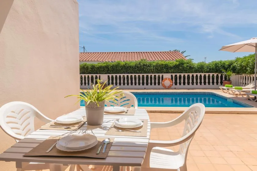 An outdoor dining table with place settings overlooks a swimming pool on a sunny terrace.