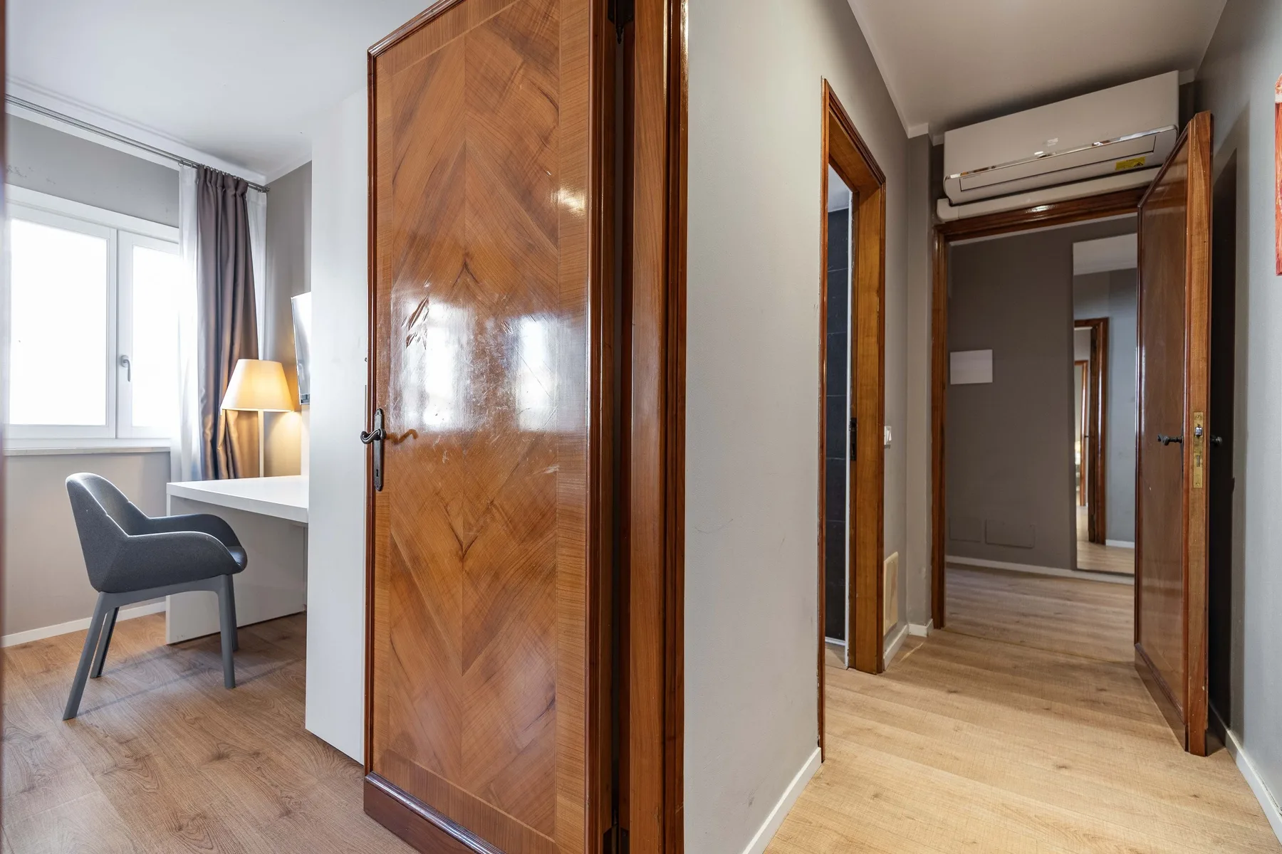 A hallway with wooden doors opens onto a small desk and chair beside a window.