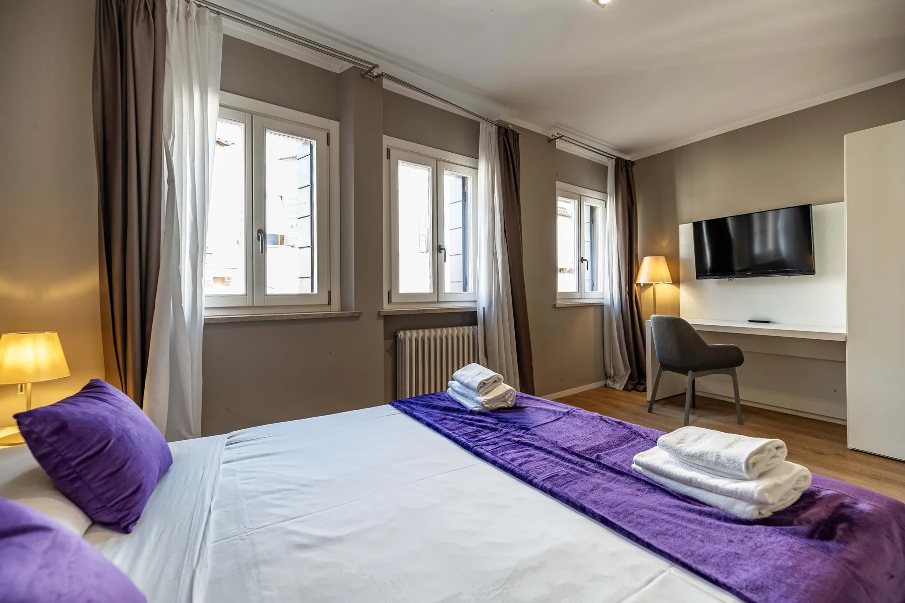 A bedroom with a bed dressed in white and purple linens beside several windows.