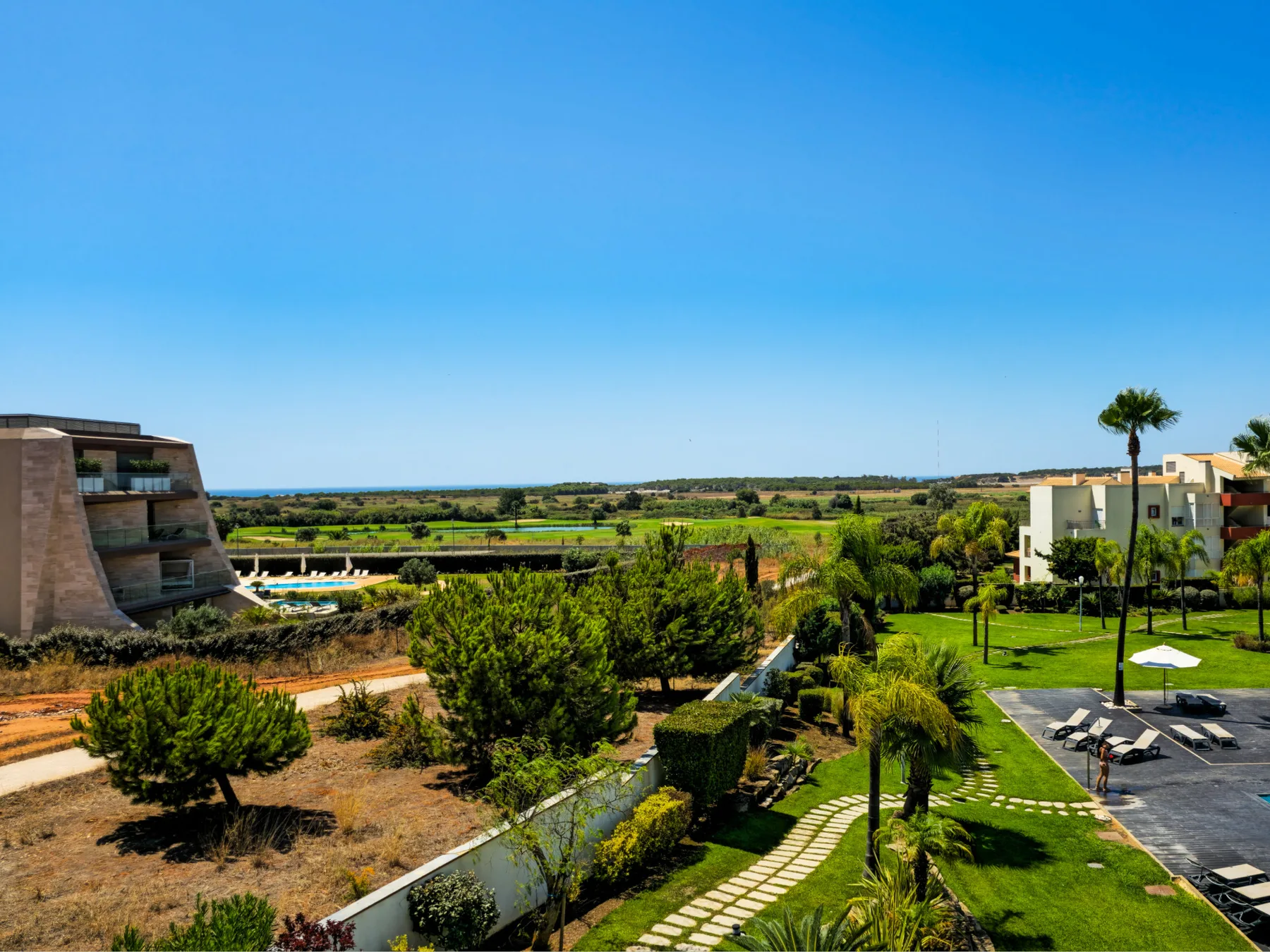 A landscaped outdoor area with palm trees, a pool, and distant green fields.