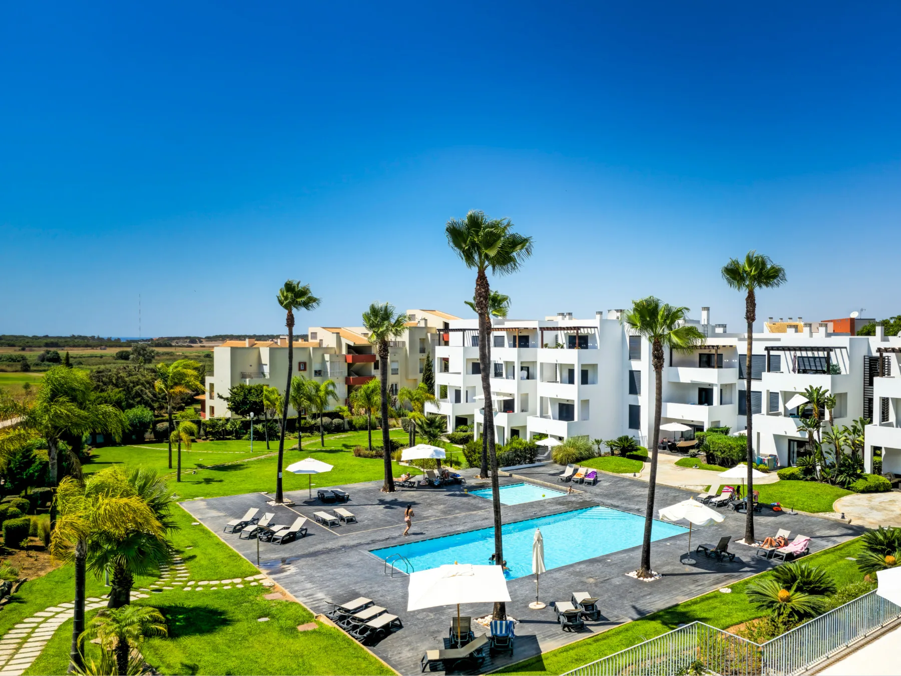 White apartment buildings overlook an outdoor pool surrounded by palm trees and greenery.