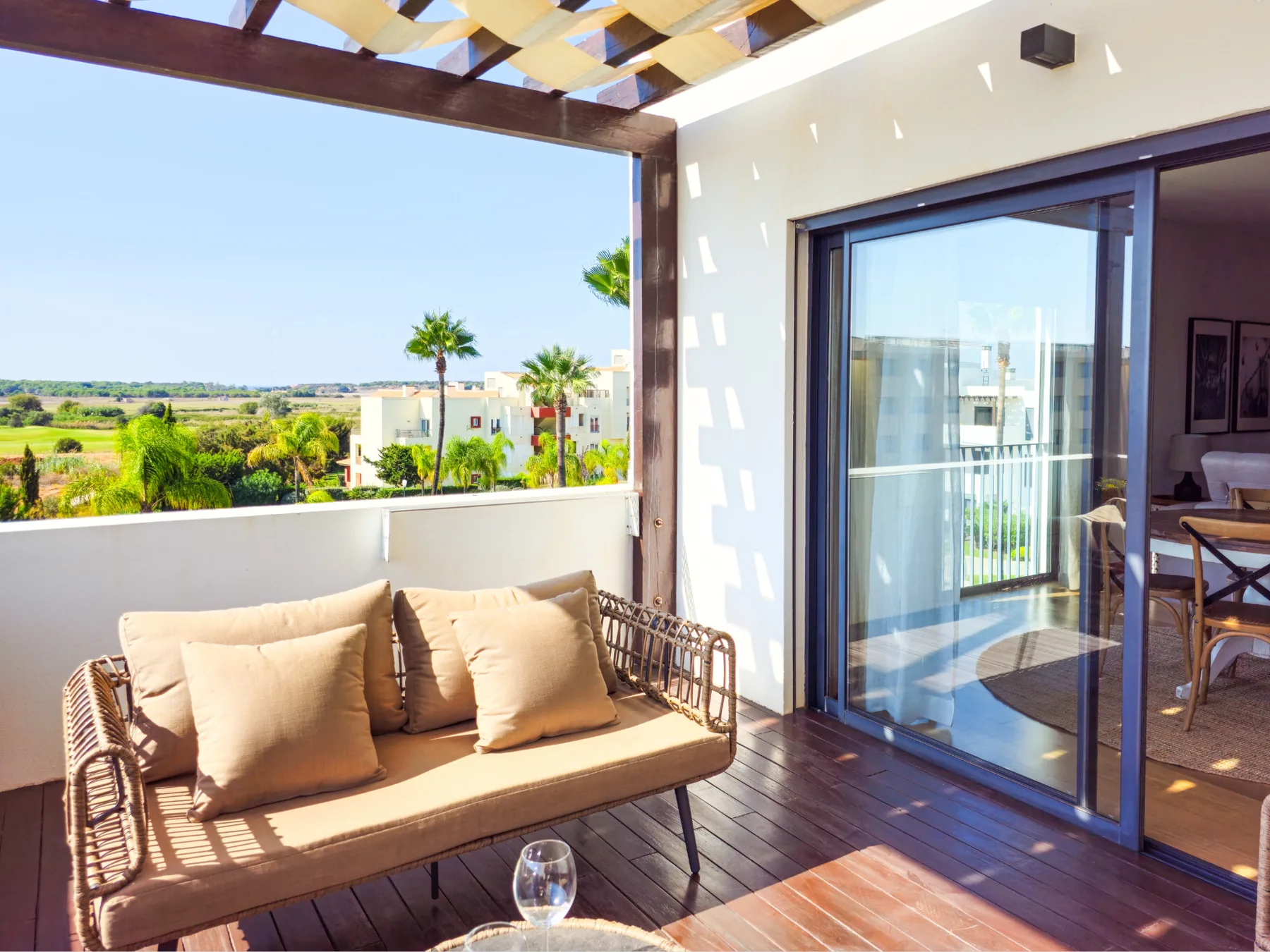 A sunny balcony has cushioned outdoor seating beneath a pergola, overlooking greenery.