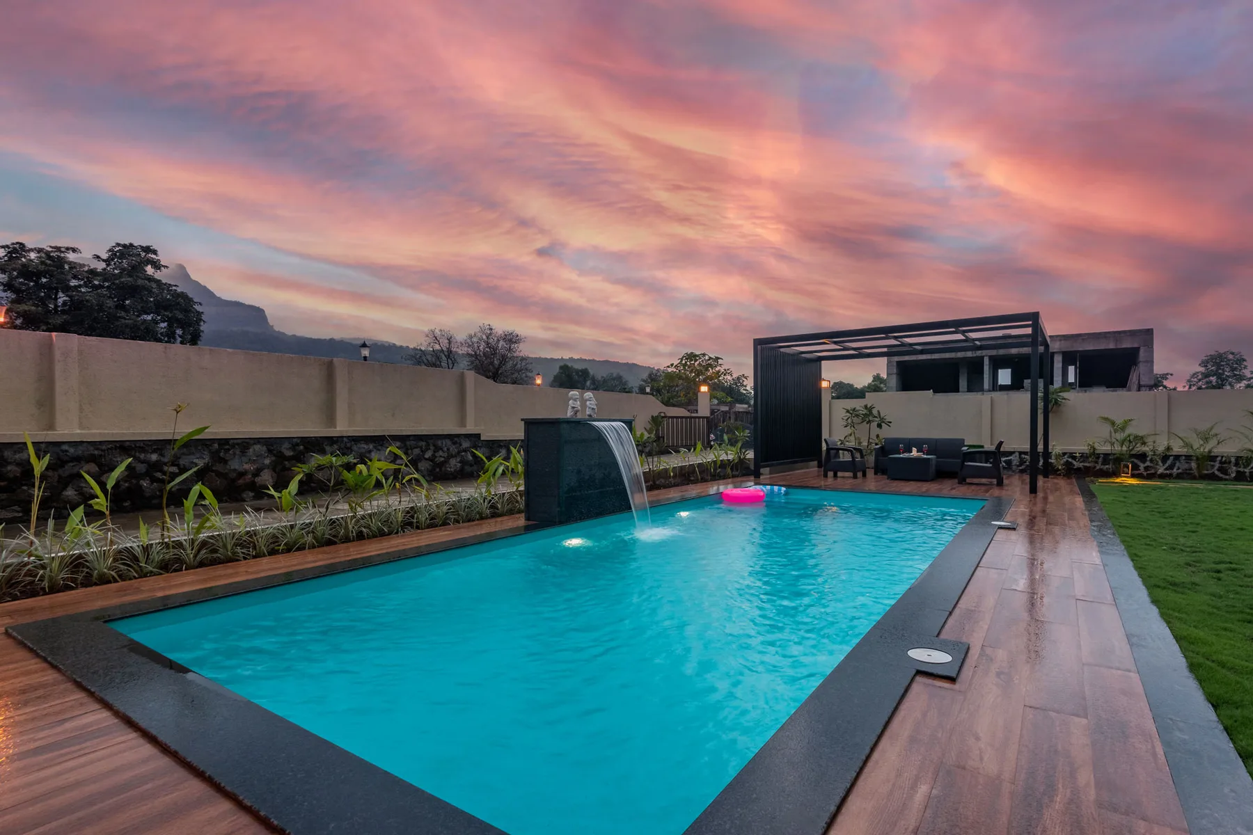 Una piscina al aire libre junto a una terraza de madera bajo un cielo colorido al atardecer.