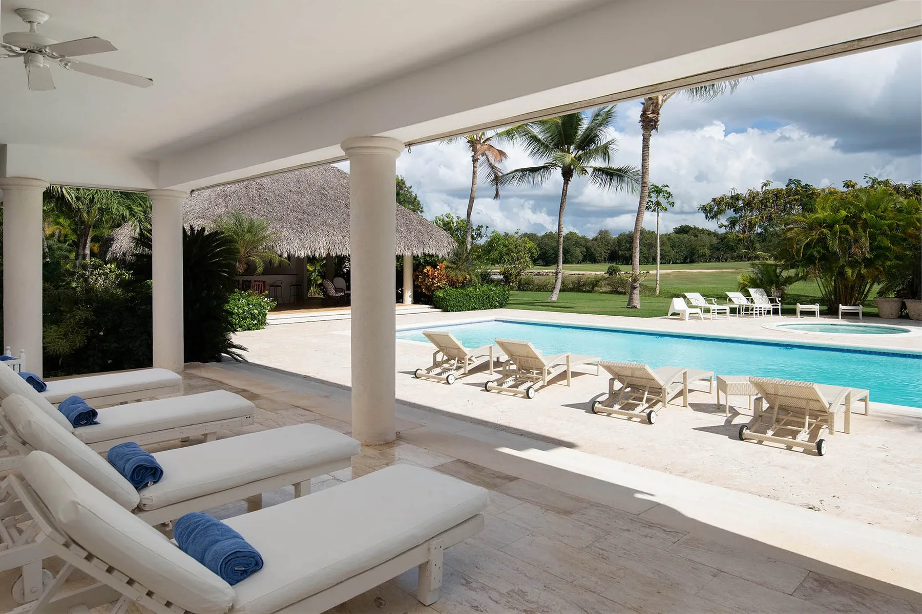 A covered patio overlooks a swimming pool with loungers and palm trees.