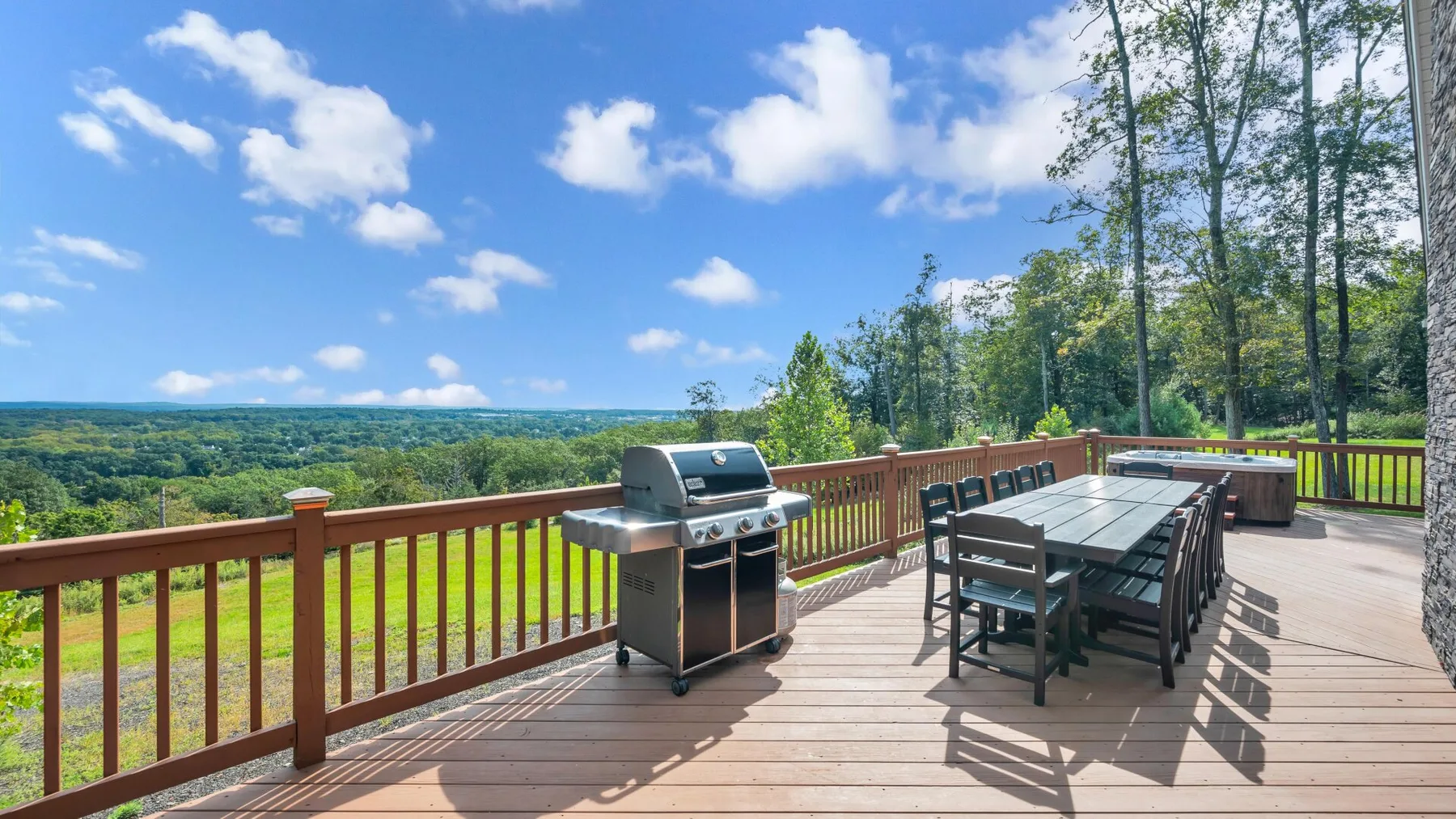 An outdoor deck with a grill and a long dining table overlooks a wooded landscape.