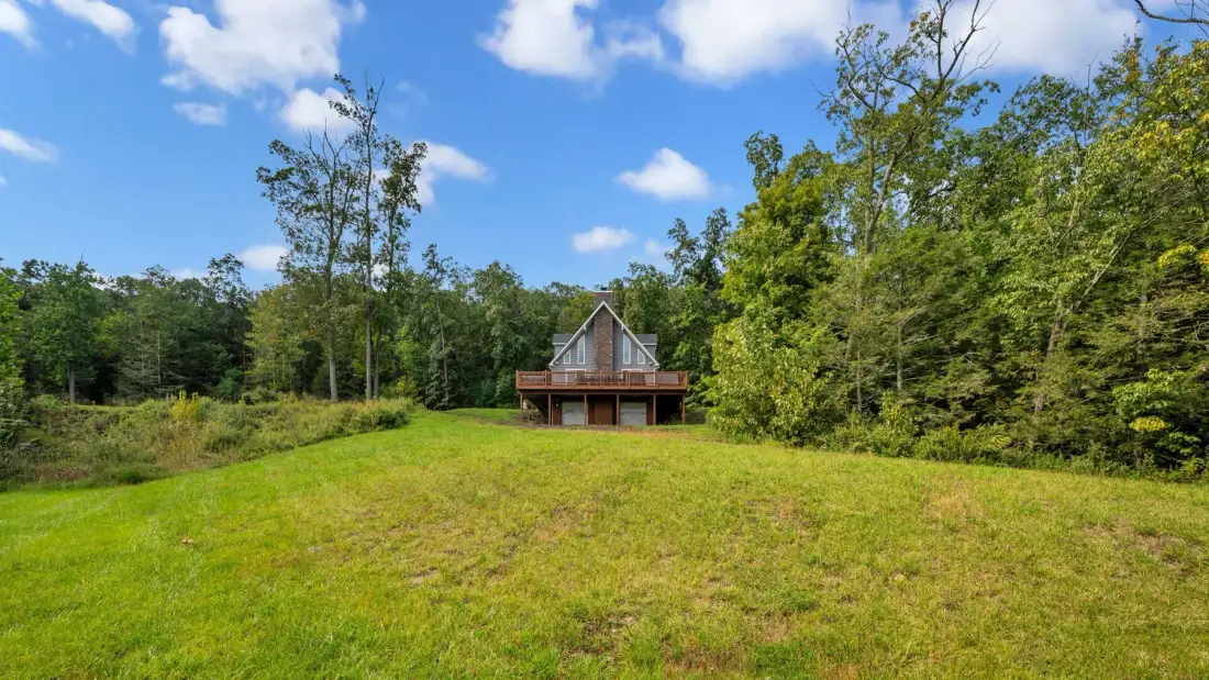 A small house stands beyond an open grassy field, surrounded by trees.