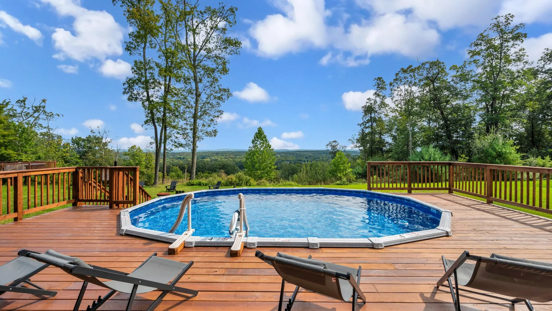 An outdoor swimming pool is set into a spacious wooden deck surrounded by trees.