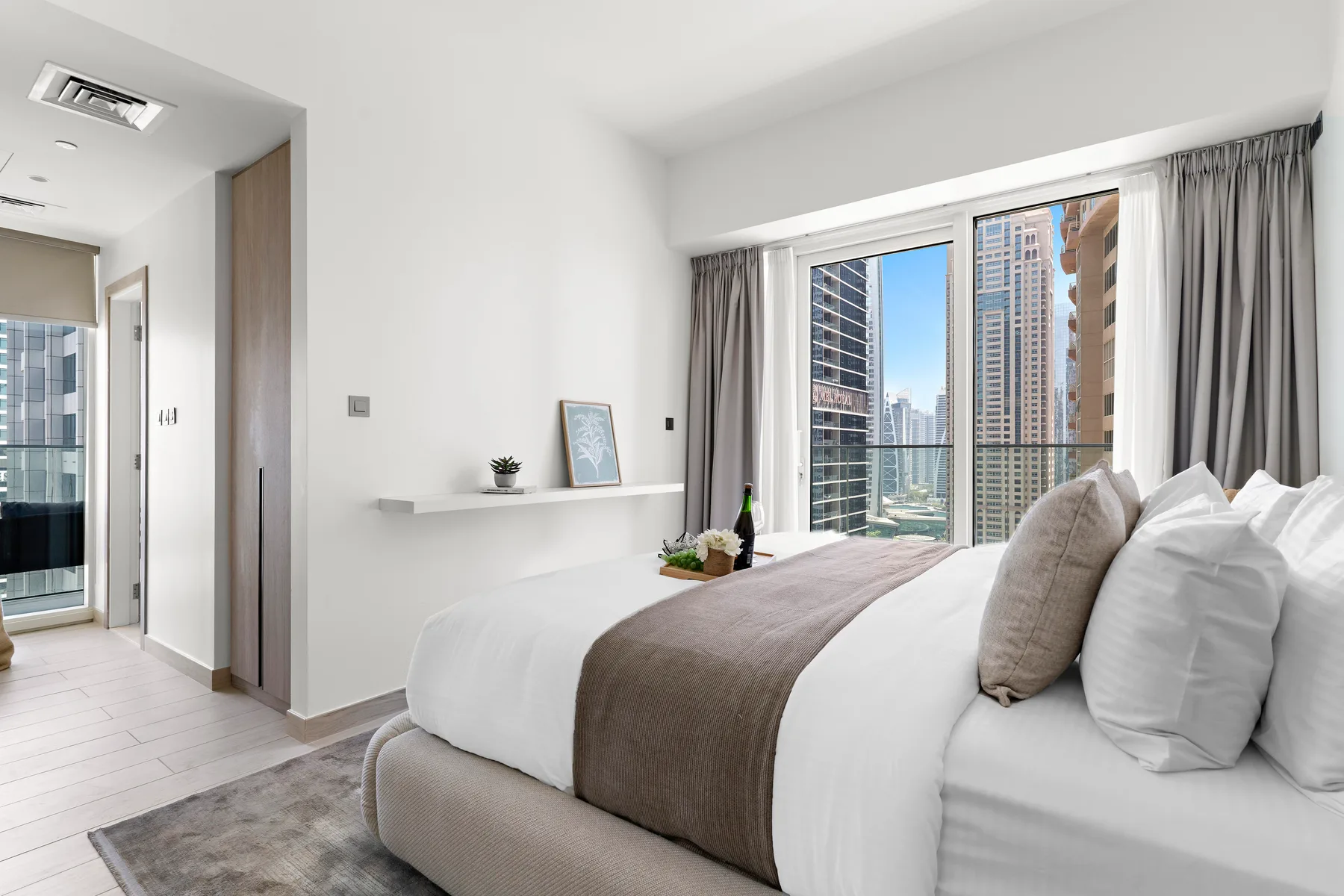 Un dormitorio luminoso con ropa de cama blanca y ventanas altas con vista a edificios cercanos.