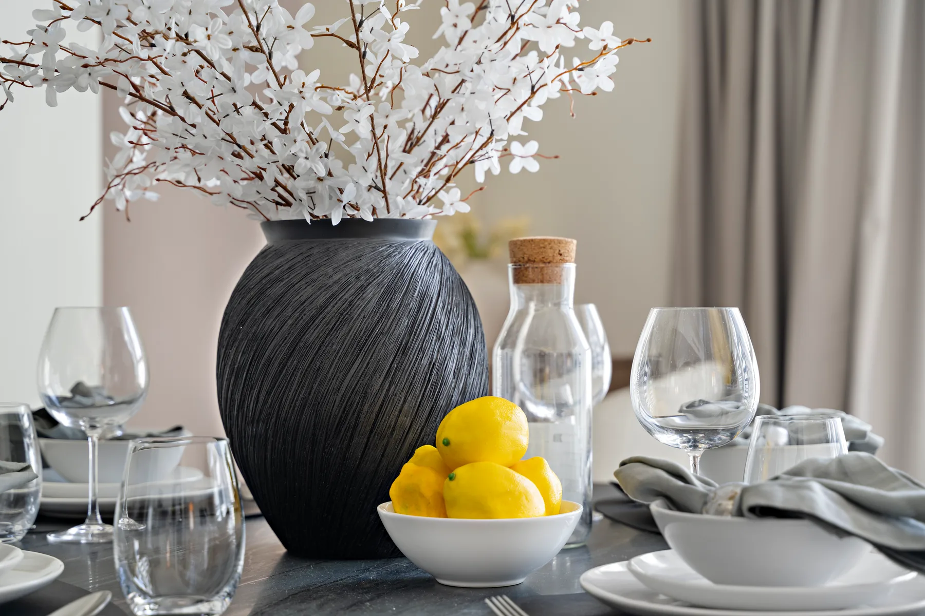 Una mesa de comedor está puesta con cristalería, platos, limones y un arreglo floral.
