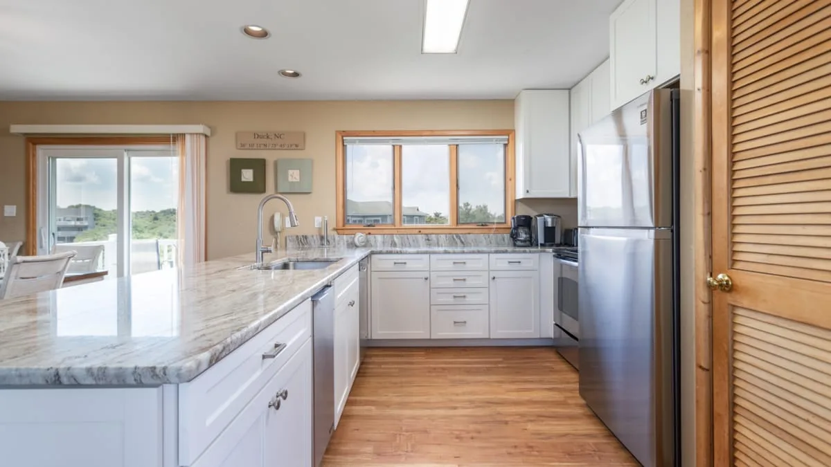 Una cocina luminosa tiene gabinetes blancos, una isla con aspecto de mármol y electrodomésticos de acero inoxidable.