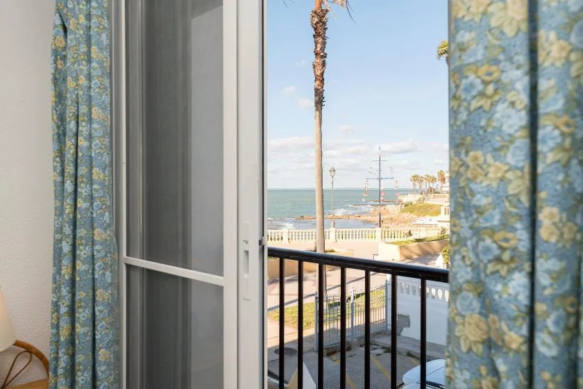 An open balcony doorway with patterned curtains overlooks the sea and promenade.