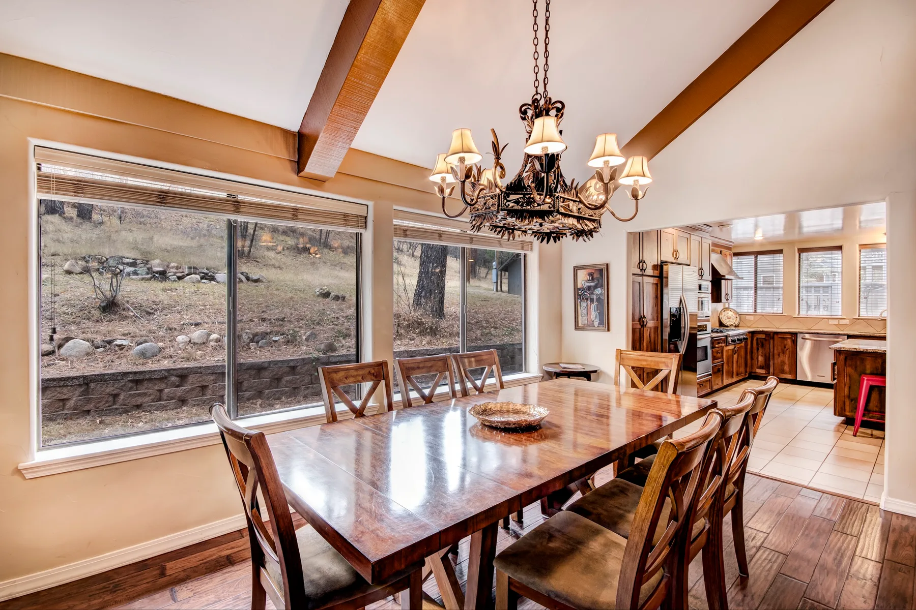 A dining room with a long wooden table and chandelier, opening onto a kitchen.