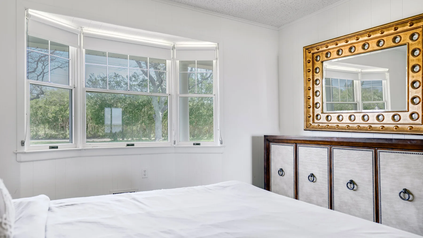 Un dormitorio con una cama bajo amplias ventanas y una cómoda con un espejo encima.
