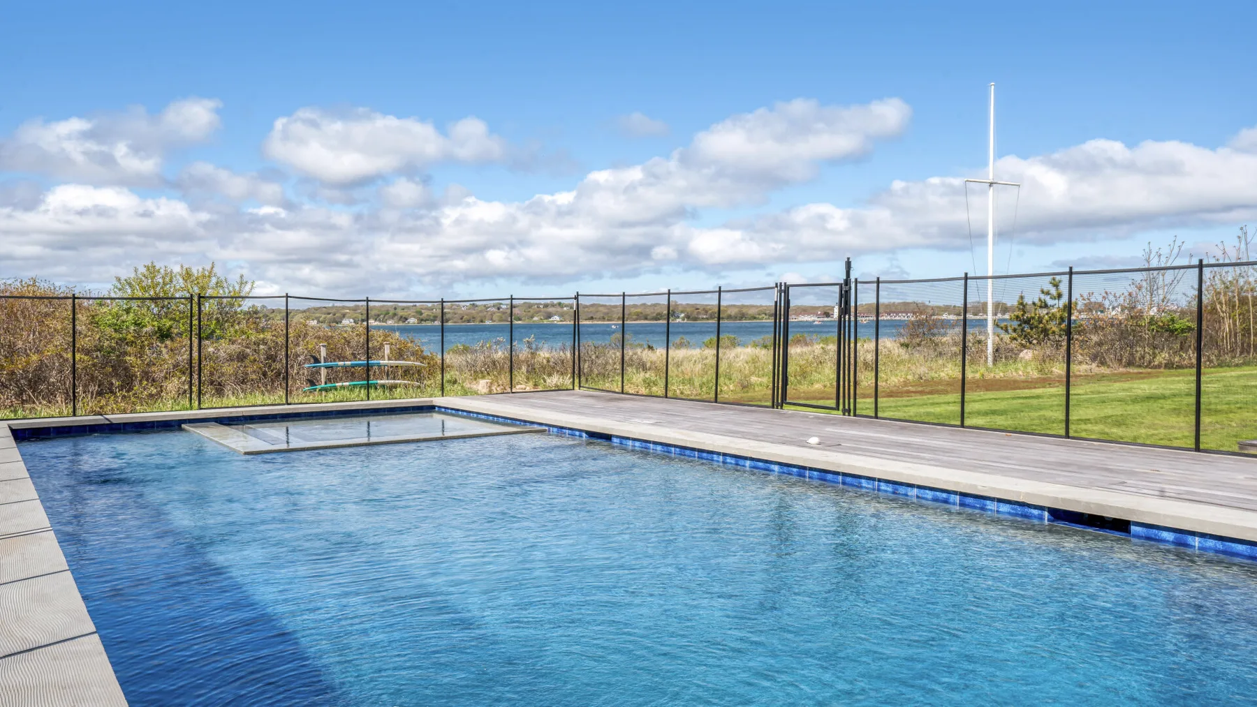 Una piscina al aire libre está junto a un césped cercado, con agua visible más allá.