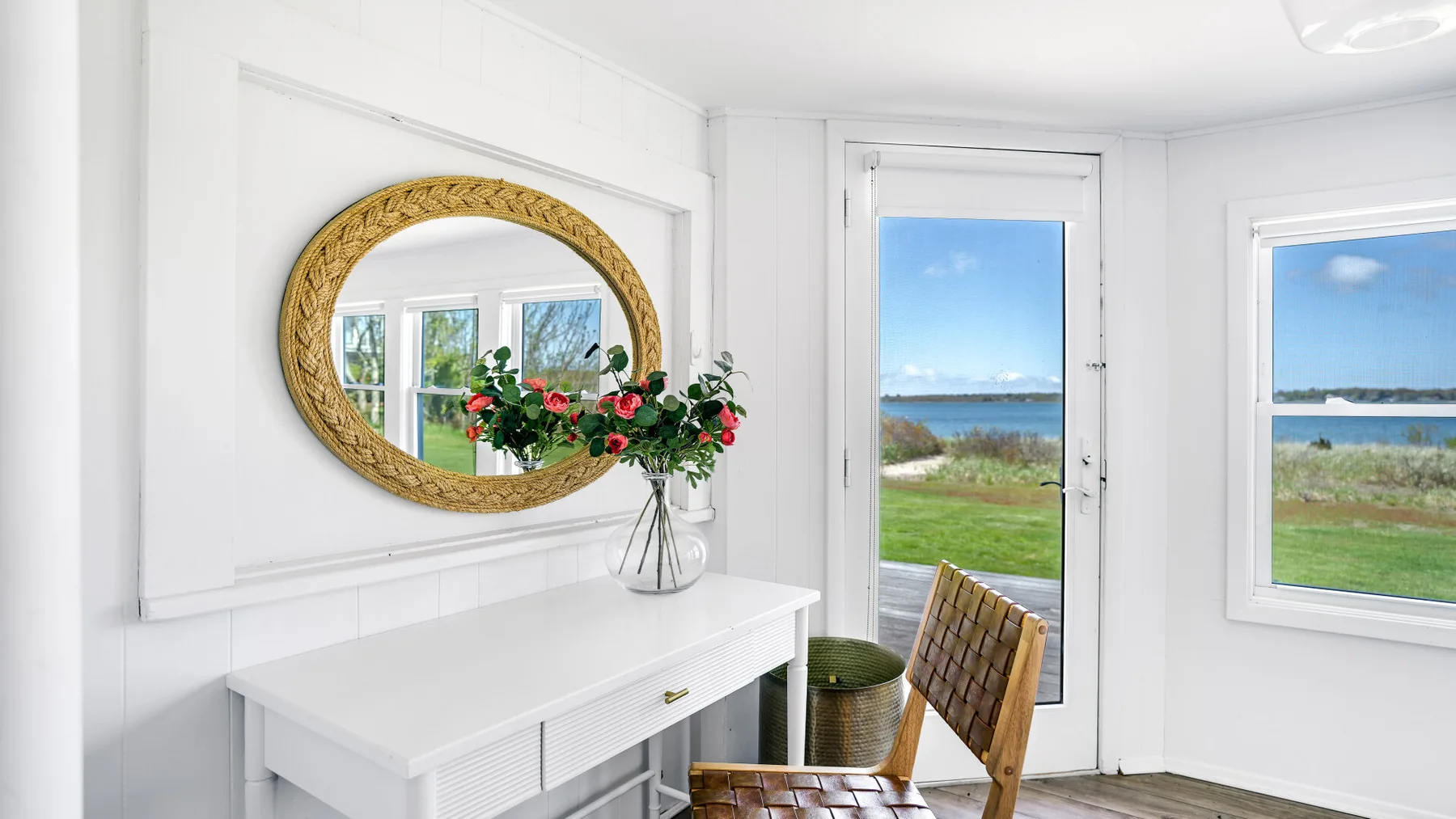 Una luminosa habitación blanca tiene una consola, un espejo ovalado y una puerta de vidrio con vista al agua.
