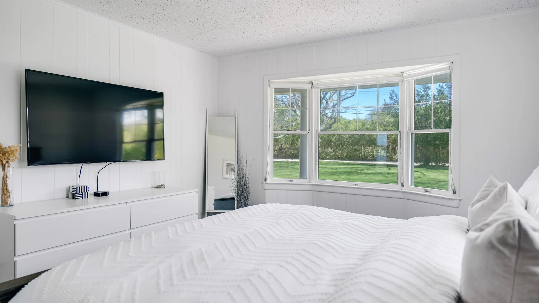 Un dormitorio luminoso con una cama grande, un televisor en la pared y ventanas con vista a la vegetación.