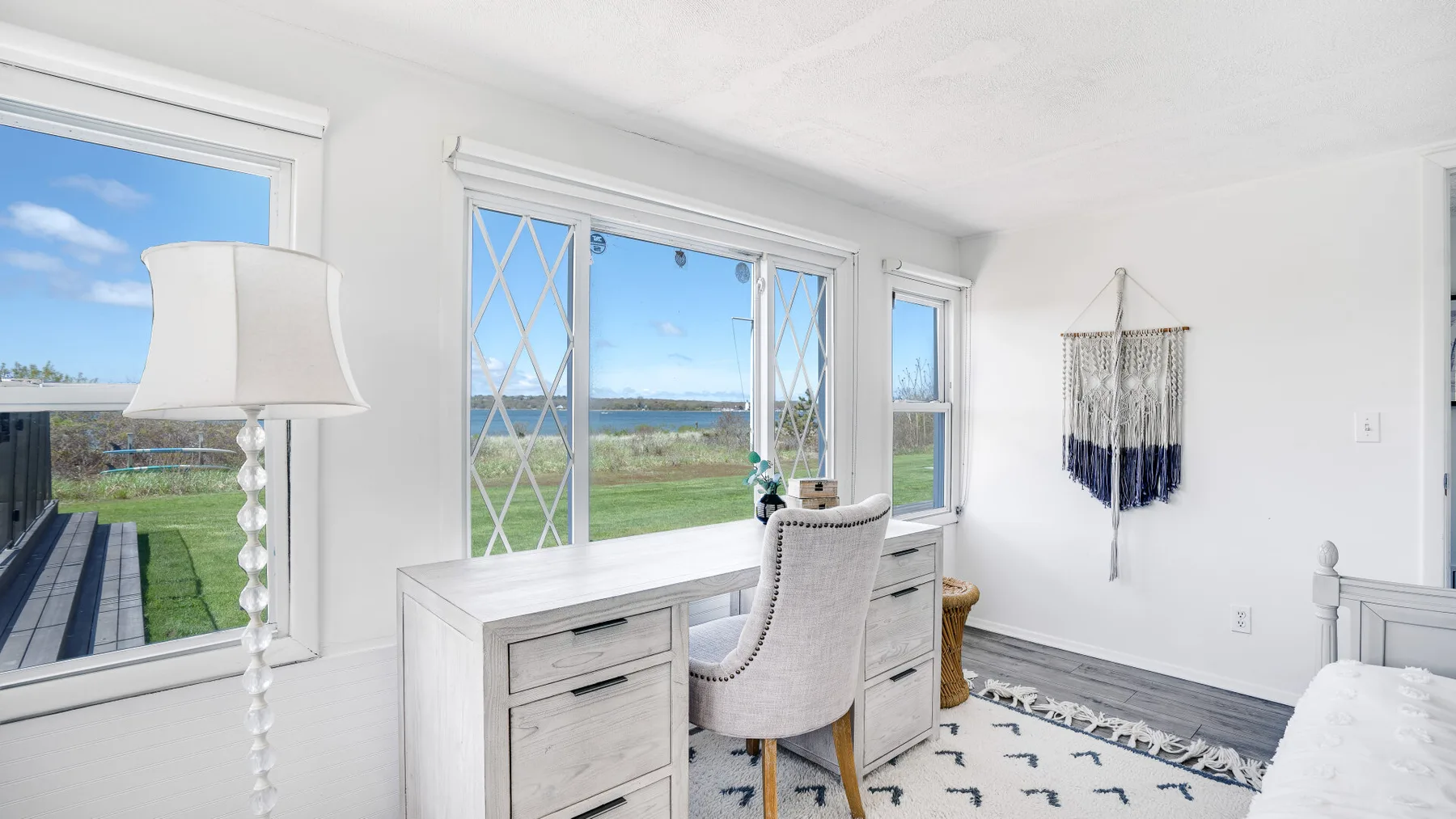 Un dormitorio luminoso con un escritorio junto a grandes ventanas con vistas a vegetación y agua.