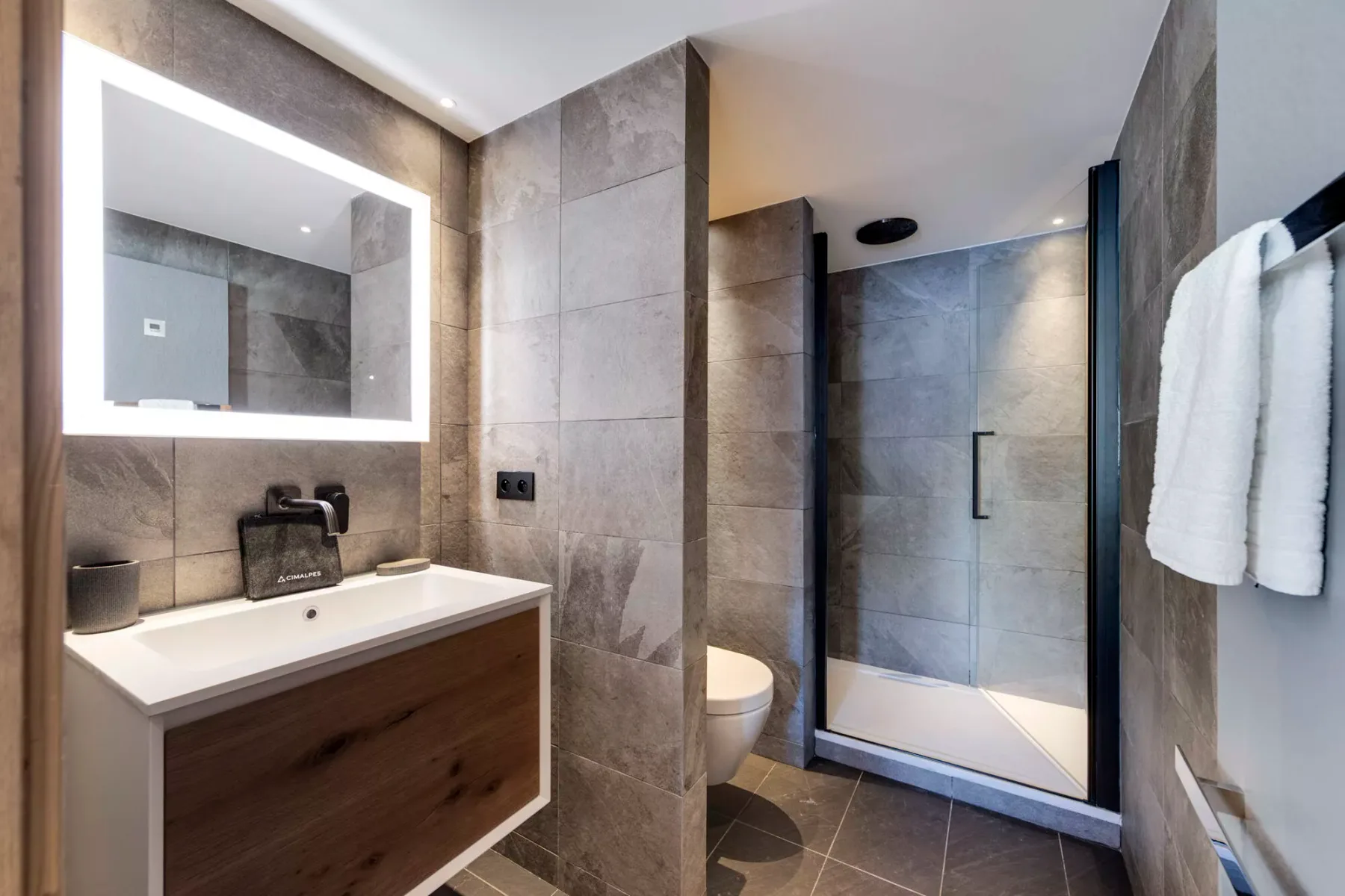 A modern bathroom with a vanity, illuminated mirror, toilet, and glass shower.