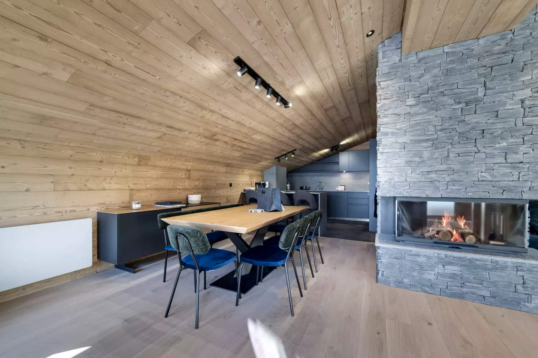 A dining area with a table beside a stone fireplace and open kitchen.