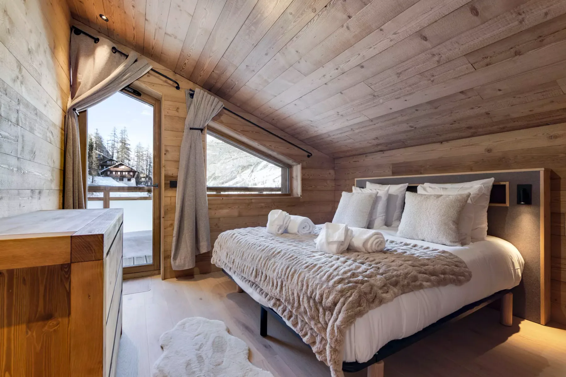 A bedroom with a bed, pale bedding, and large windows overlooking a snowy landscape.