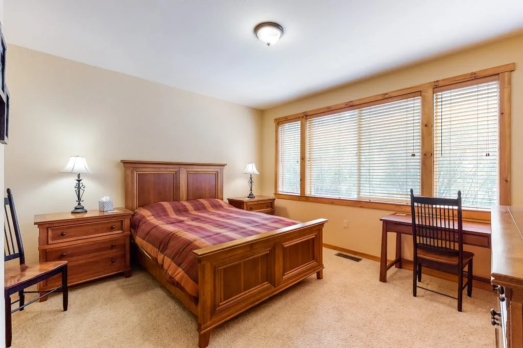 A bedroom with a wooden bed and large windows with blinds.