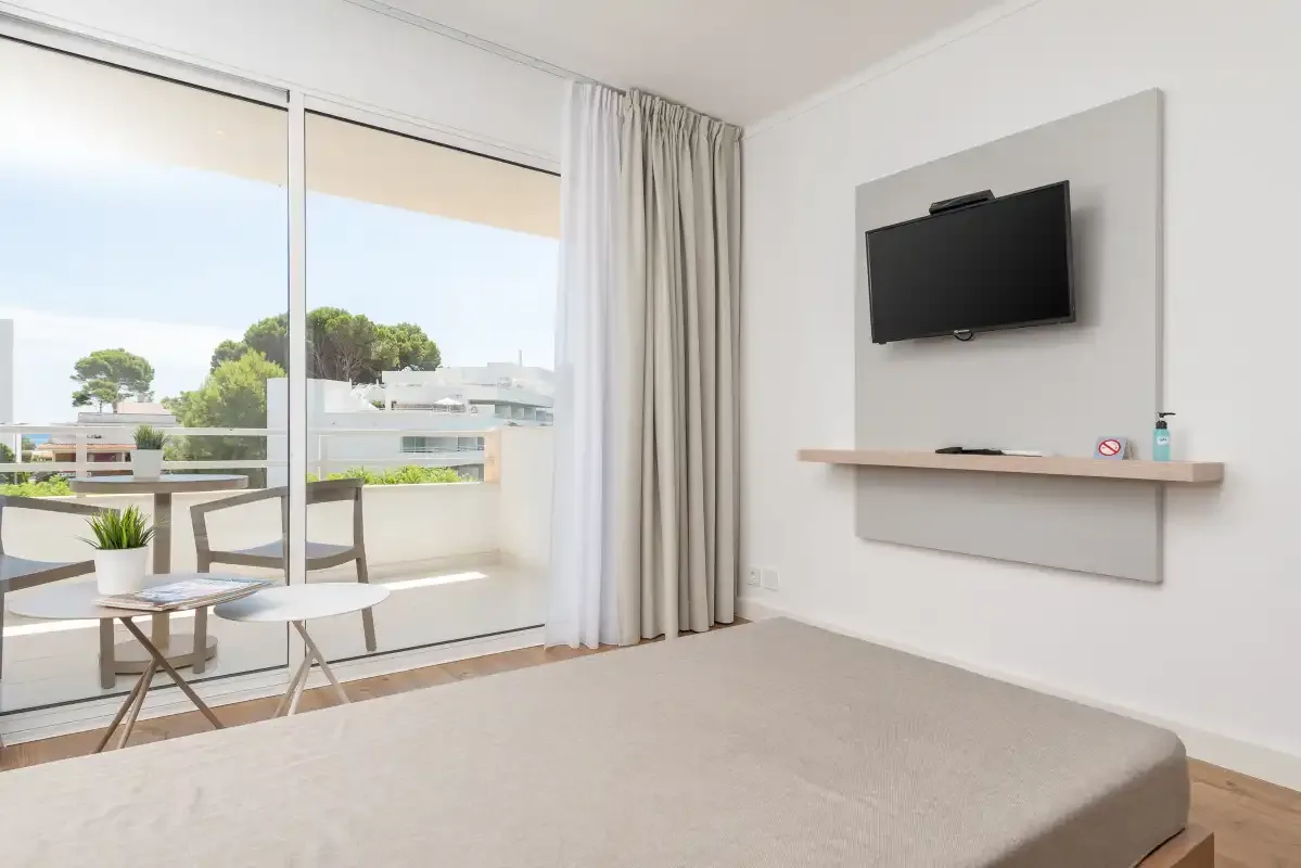 A bright bedroom with a bed, a wall-mounted television, and sliding glass doors to a balcony.