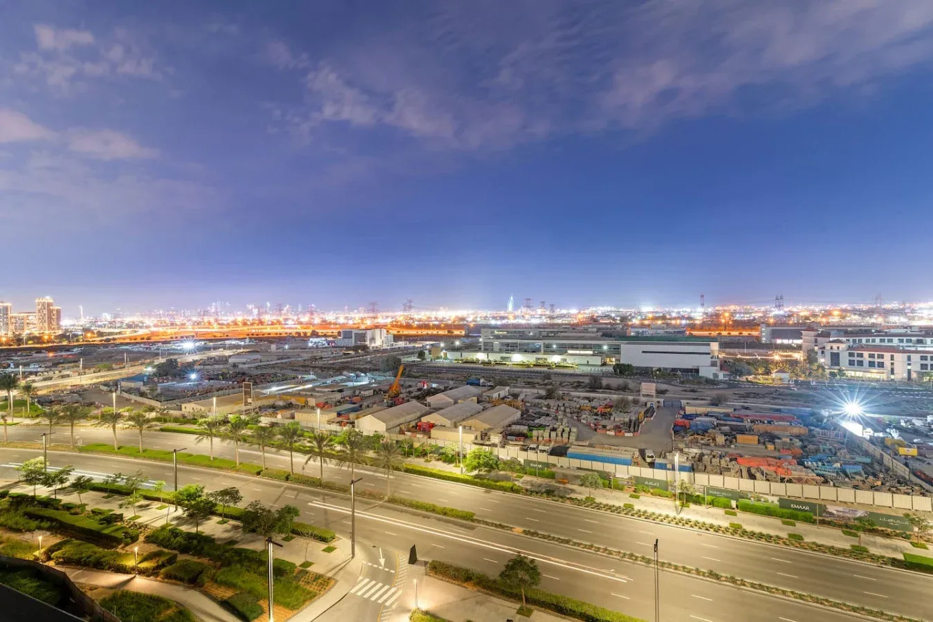 A broad road and industrial buildings stretch across a brightly lit city at dusk.