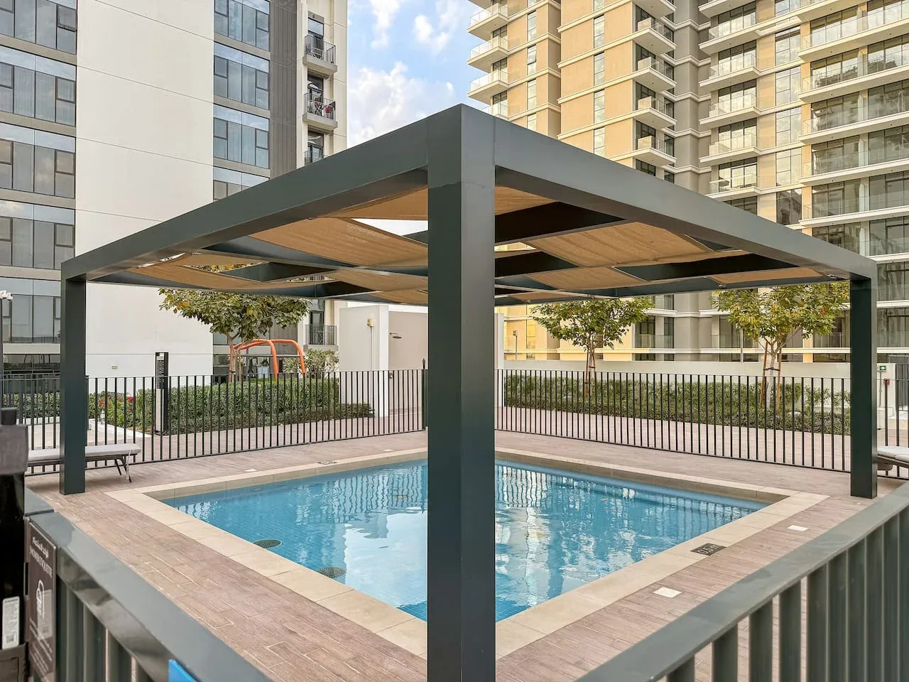 An outdoor swimming pool sits beneath a shaded pergola among apartment buildings.