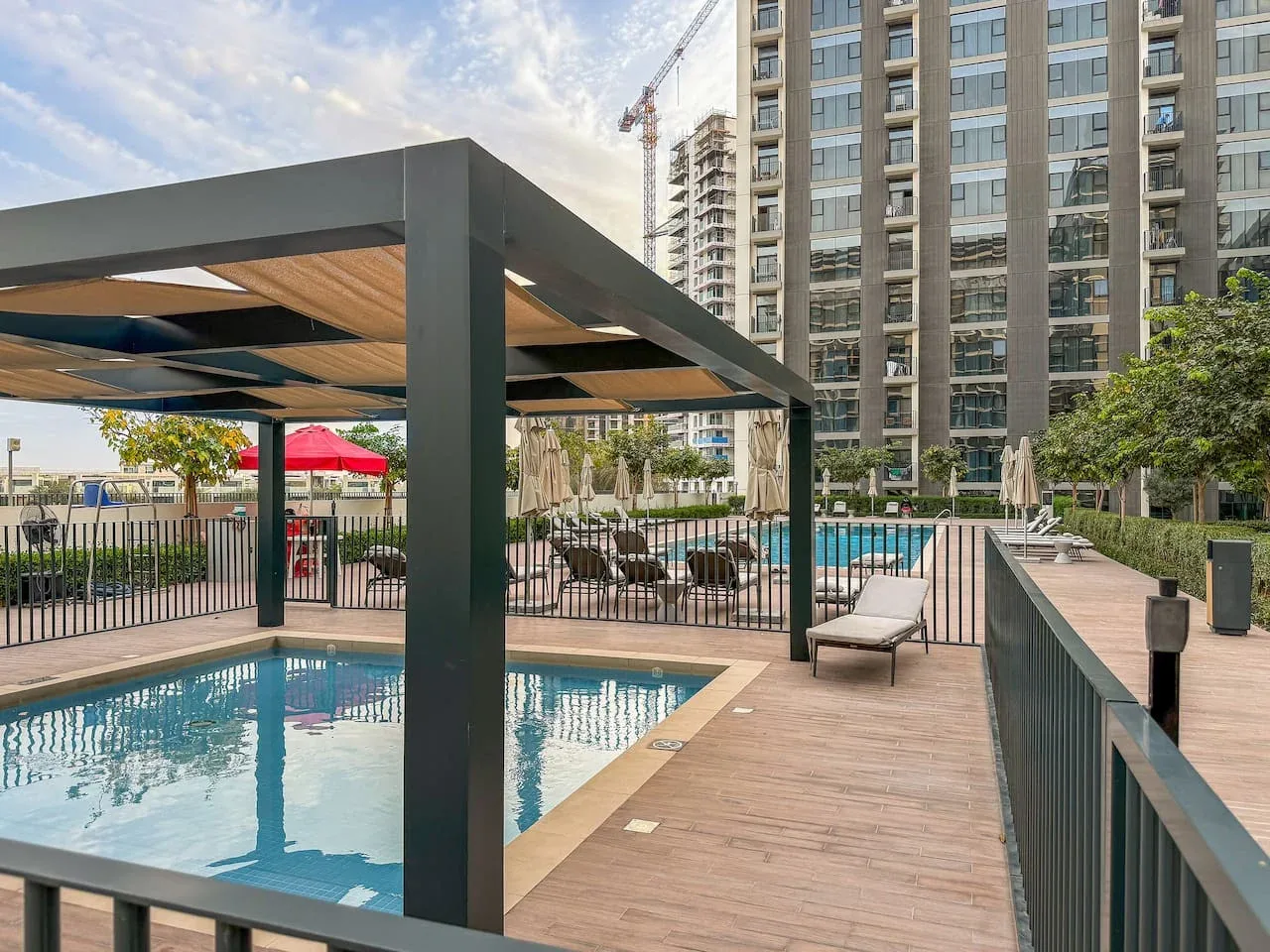 An outdoor pool deck with seating beneath a shaded pergola beside a high-rise building.