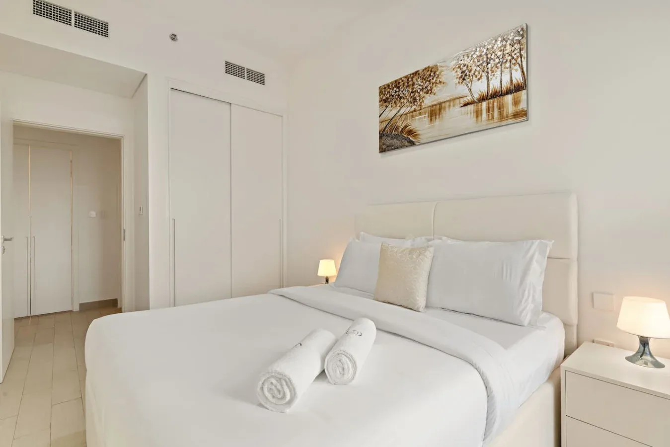 A bright bedroom with a neatly made bed and a landscape print above the headboard.