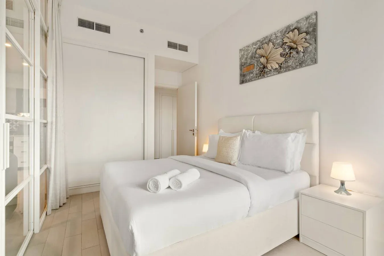 A bright bedroom with a neatly made bed and floral wall art.