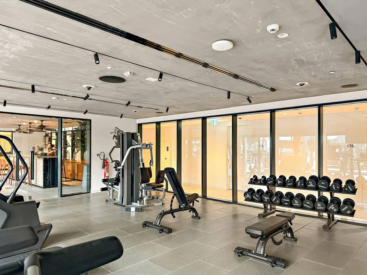 A gym with weight machines, benches, and dumbbell racks beside large windows.