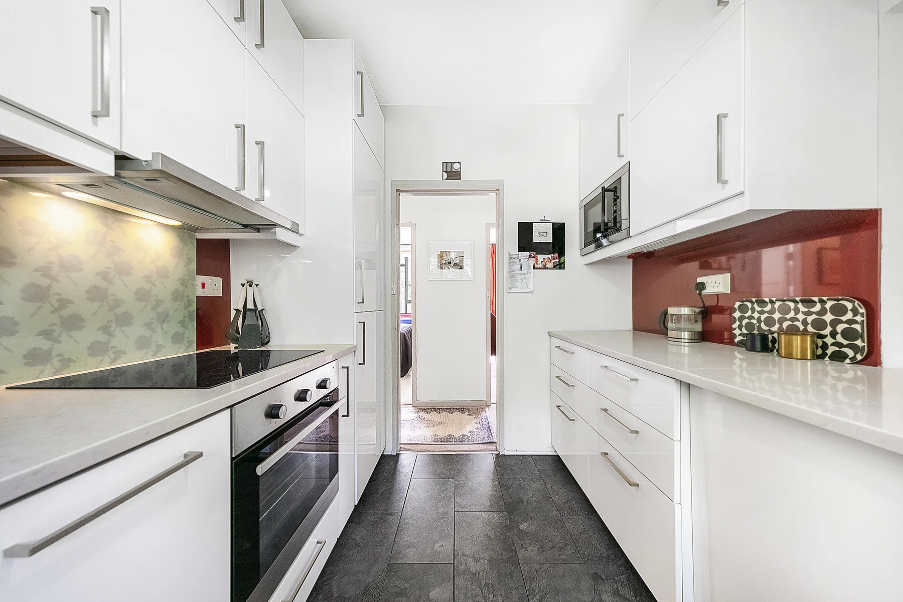 A bright galley kitchen with white cabinets, dark countertops, and a central walkway.