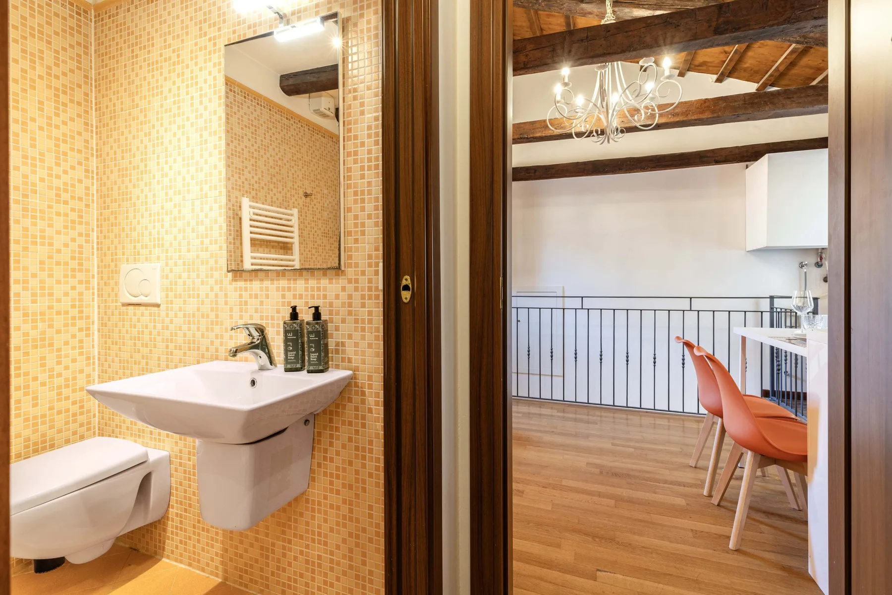 Un baño con azulejos y lavabo mural se abre a una estancia luminosa con vigas vistas.