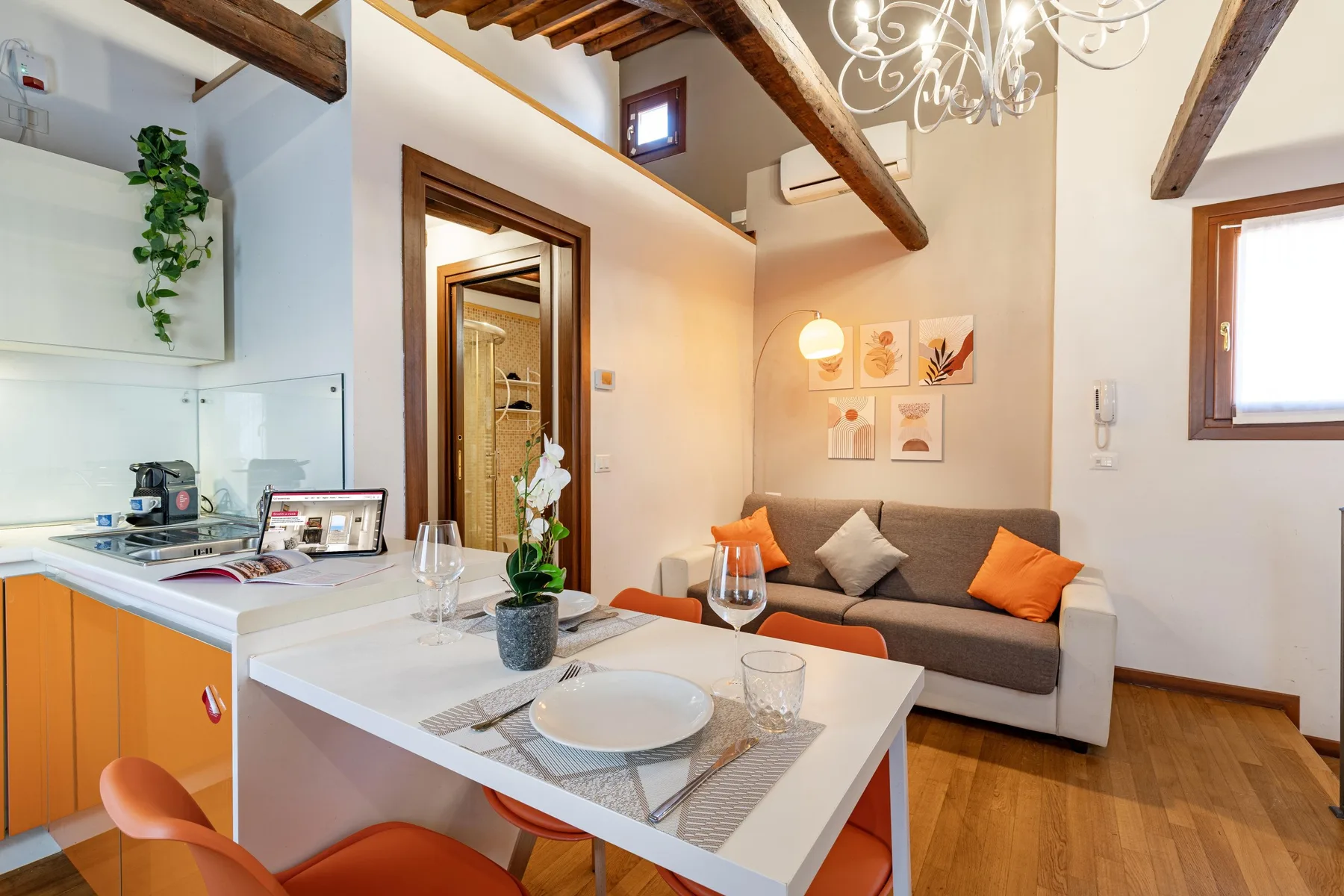 Comedor con la mesa puesta junto a una pequeña cocina y un sofá gris.