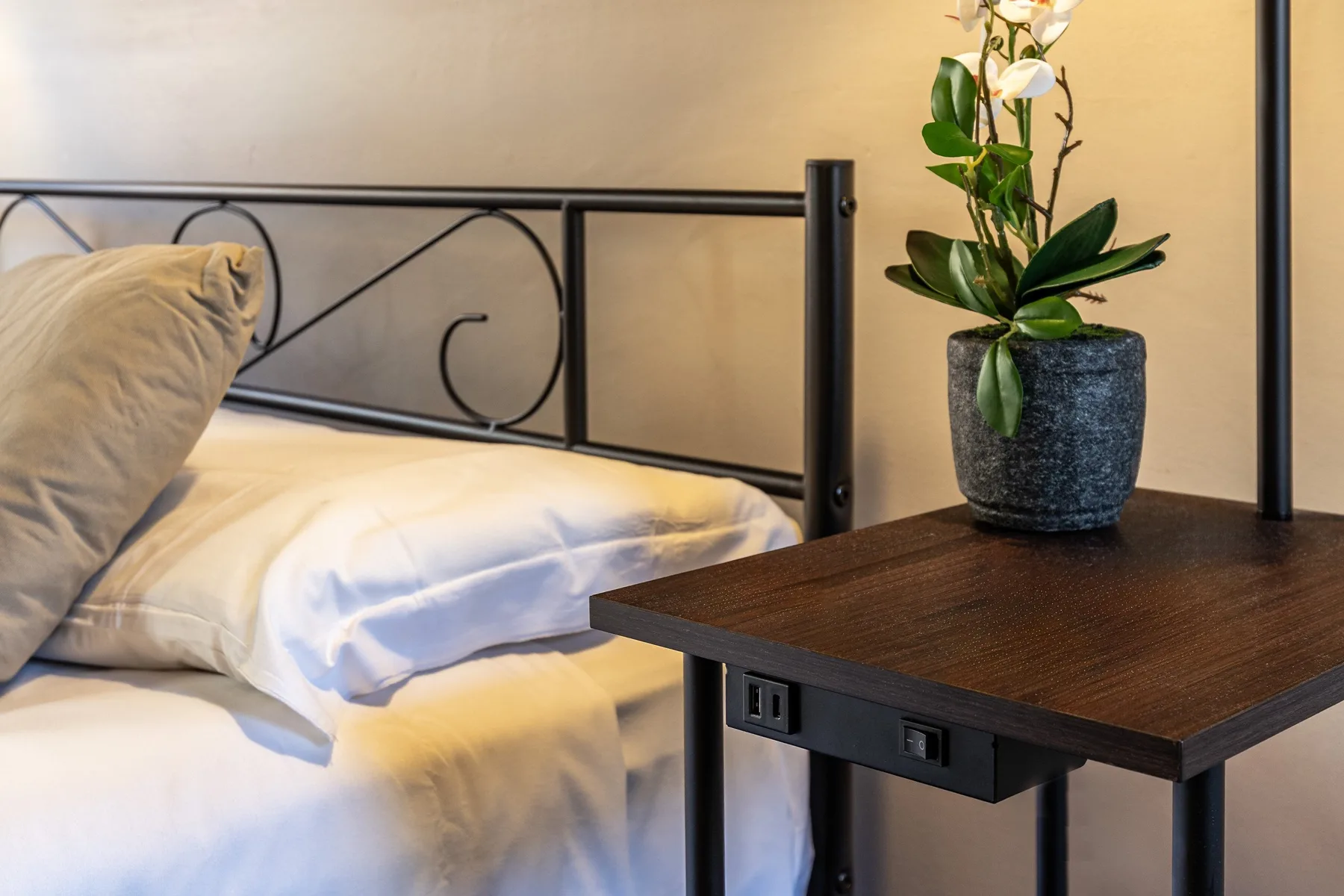 Una cama con ropa de cama clara está junto a una mesa oscura con una planta con flores.
