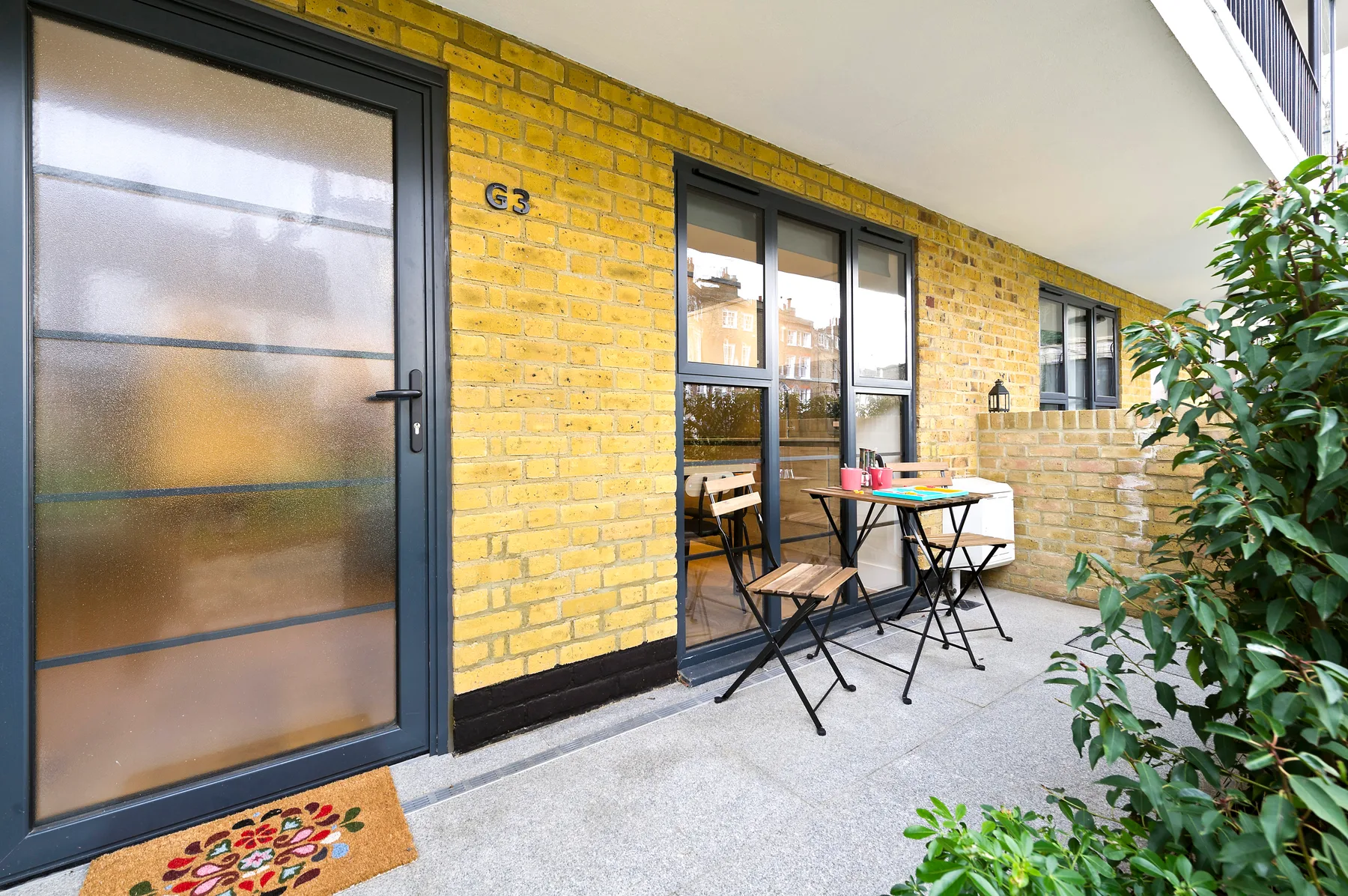 A small outdoor seating area sits beside a yellow brick wall and glass doors.
