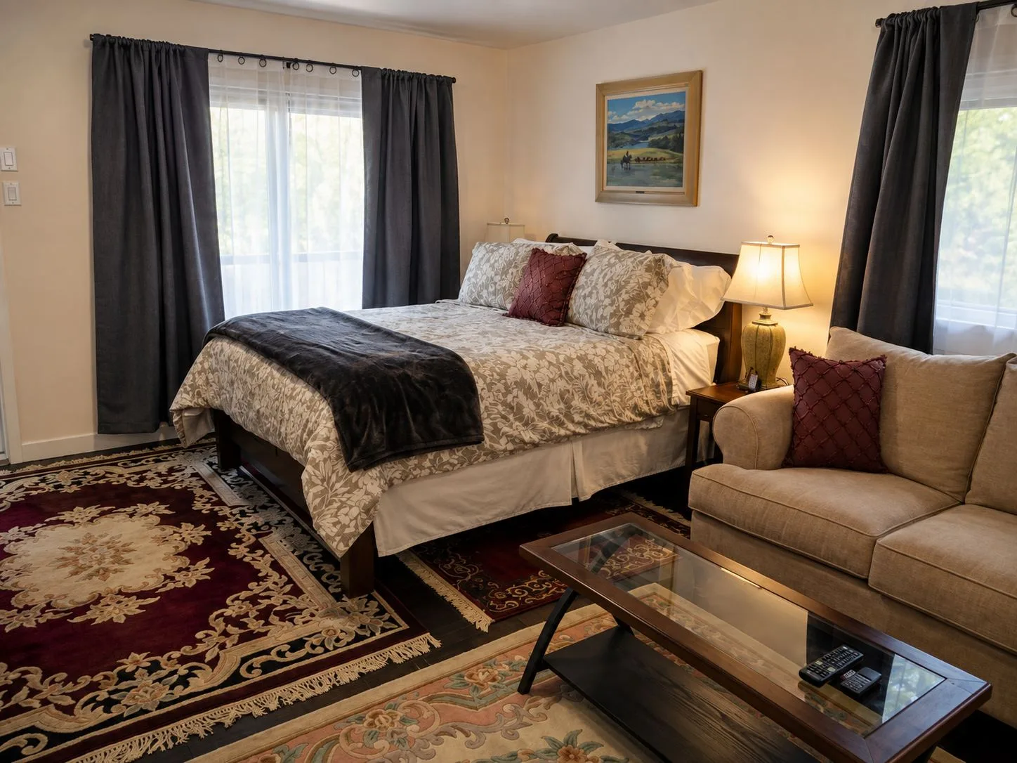 La suite Chilcotin: una cama queen con una manta oscura, un sofá y una mesa de centro junto a ella, y luz natural que entra por ventanas con cortinas en dos lados.