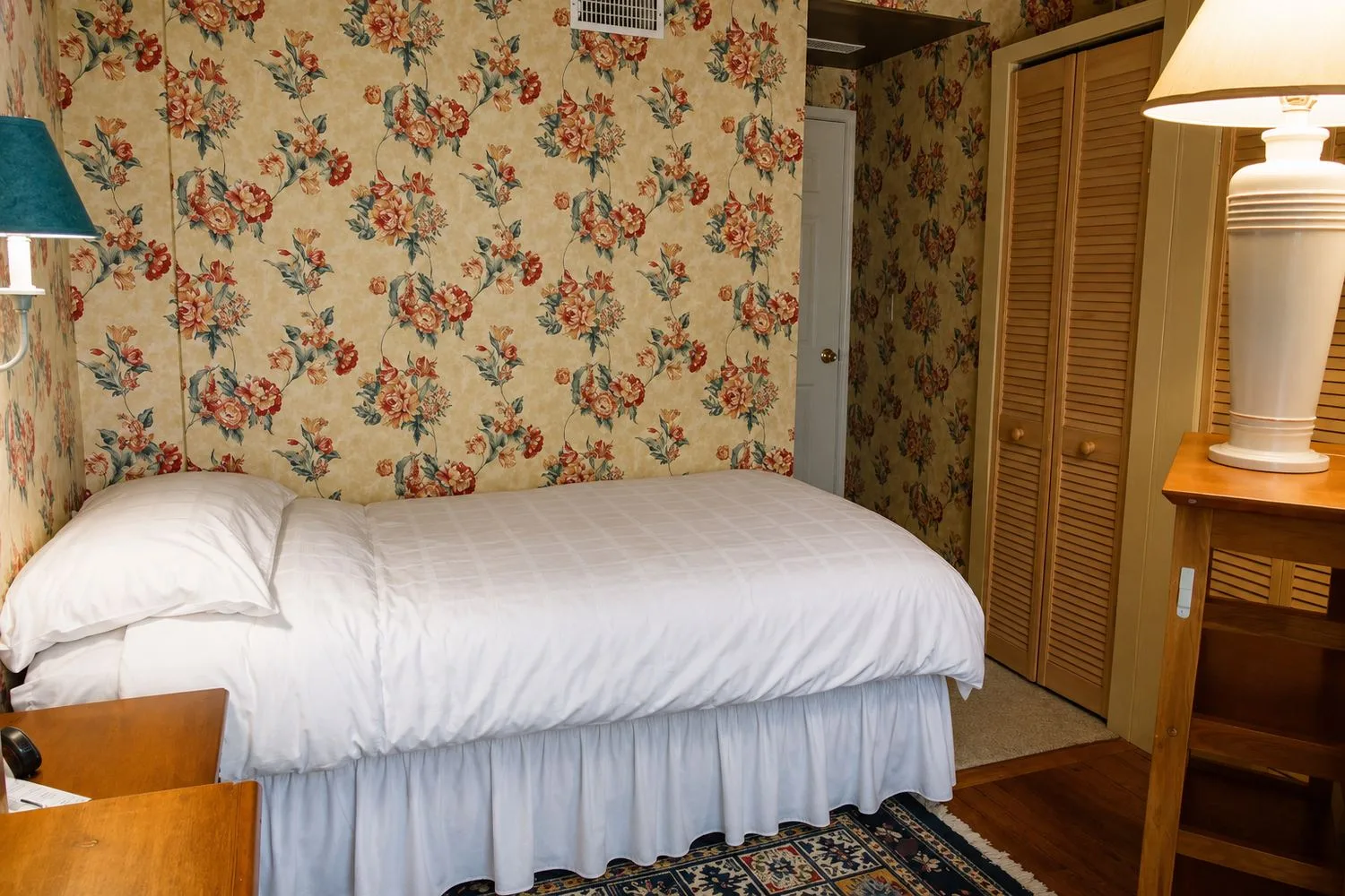 La habitación Pelican: una cama vestida de blanco junto a paredes con papel tapiz de flores, con un armario de puertas de lamas y una lámpara sobre la mesa auxiliar.