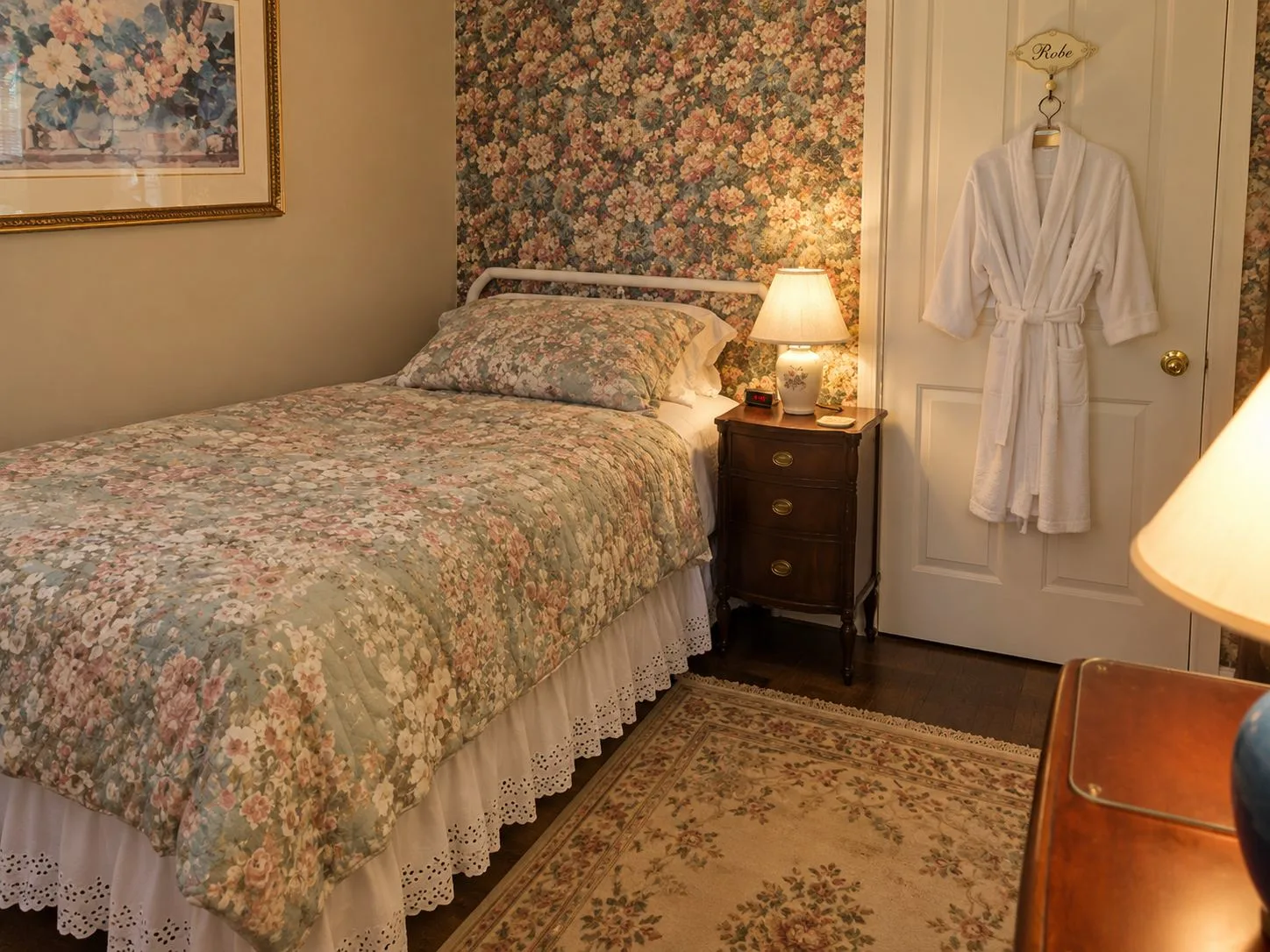 La habitación Pine: una cama individual con una colcha de flores junto a una pared con papel tapiz estampado, una lámpara encendida sobre la mesa de noche y una bata colgada en la puerta.