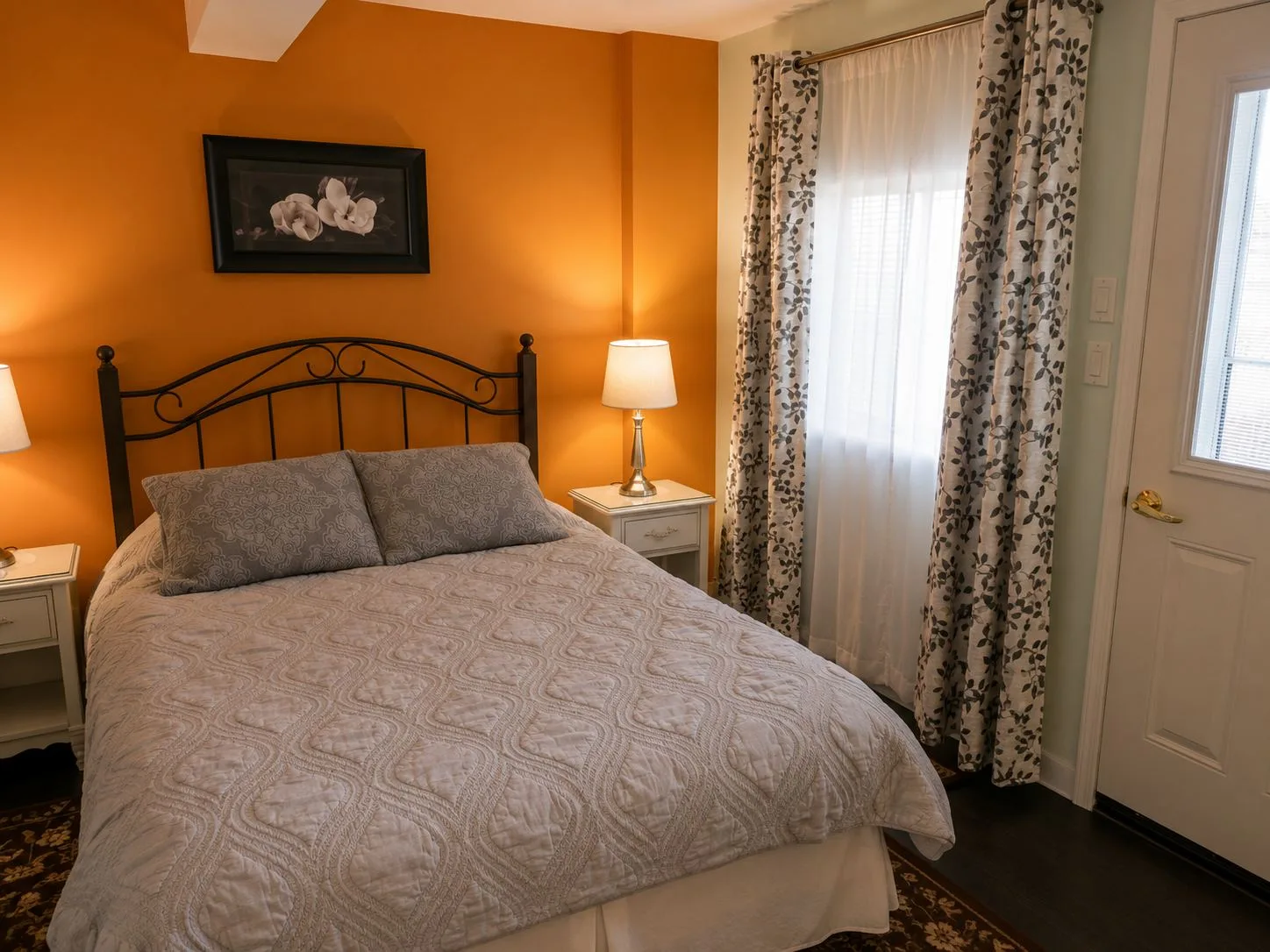 La suite Blue Heron: una cama queen junto a una pared de un cálido tono naranja, una lámpara sobre la mesa de noche y una puerta con cortina que da al exterior.