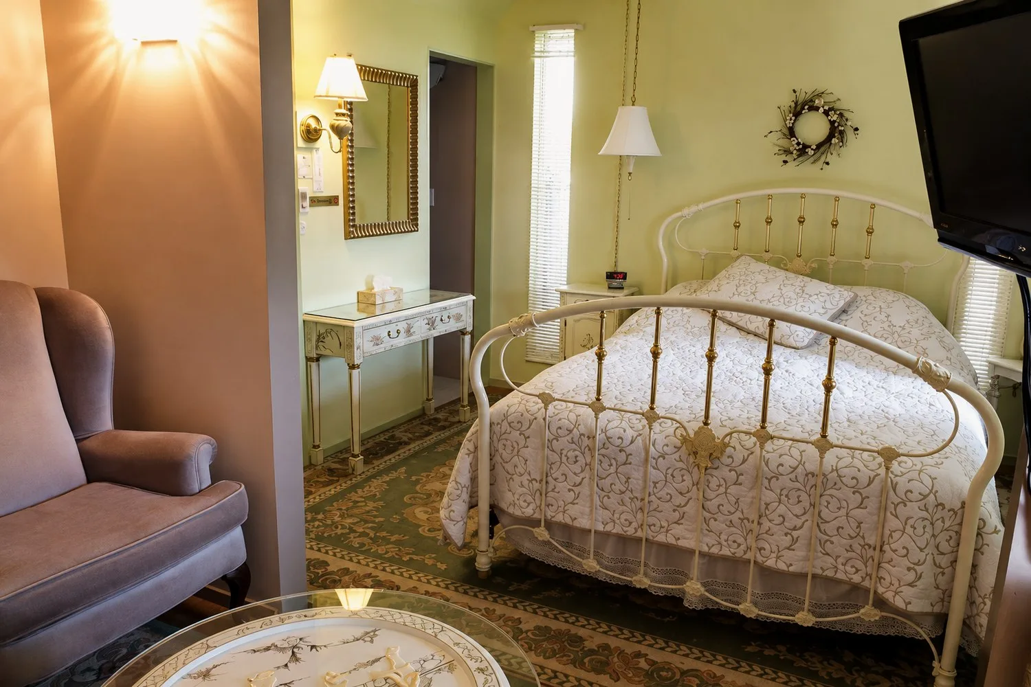 La suite Green: una cama queen con estructura blanca y de latón y una colcha color crema, paredes verde claro y un sillón en primer plano.
