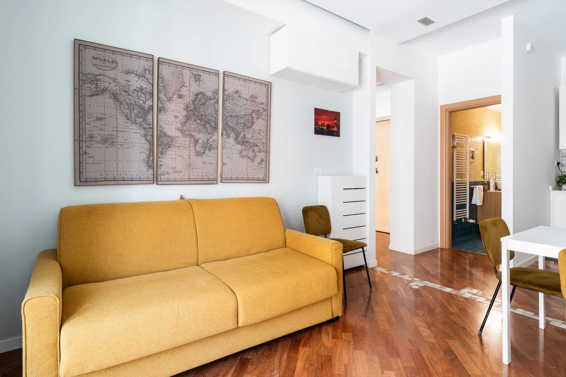 A bright living area with a yellow sofa, framed maps, and a wooden floor.