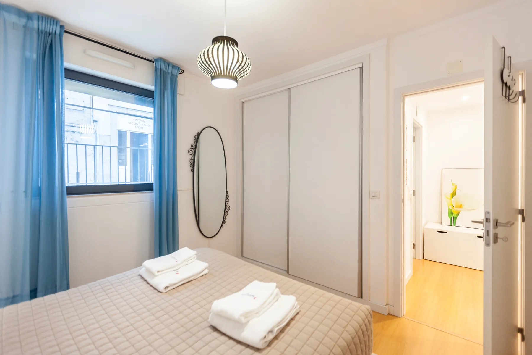Dormitorio luminoso con una cama, cortinas azules y una puerta hacia otra habitación.